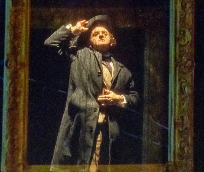 A man standing behind a frame, wearing a long gray coat and lifting off his large hat.